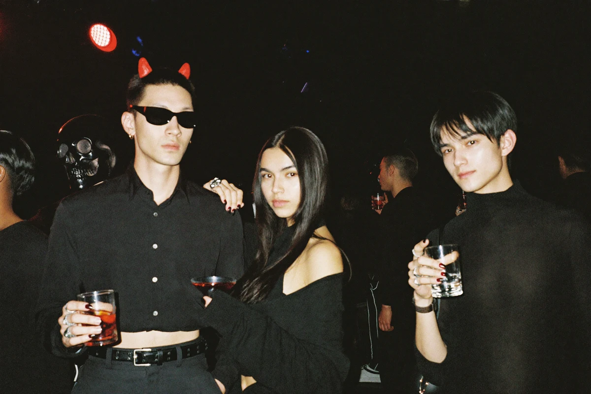 Young adults in chic Halloween costumes holding cocktails at a neon-lit nightclub bar crawl