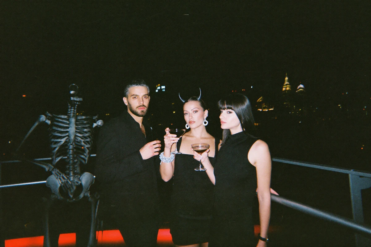 Friends in minimalist Halloween costumes clinking cocktails at a rooftop bar with a city skyline at night
