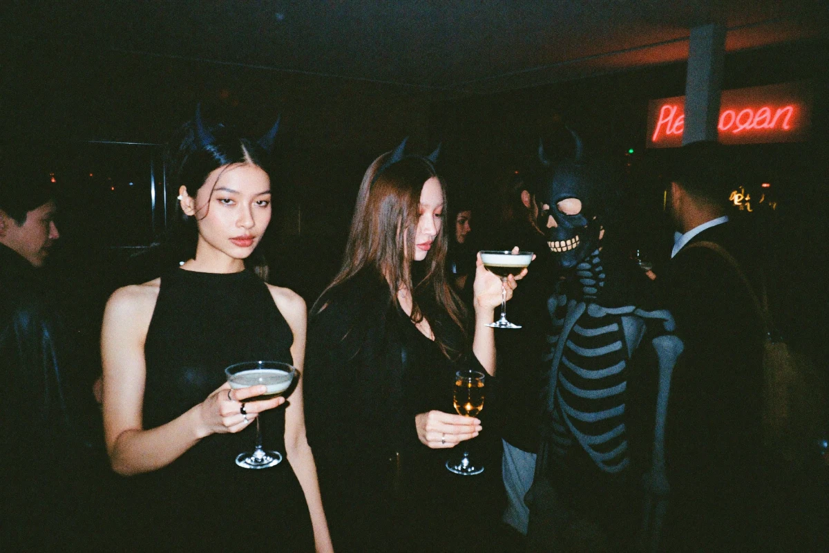 Young adults in minimalist Halloween costumes clinking cocktails at a crowded speakeasy bar crawl under red neon