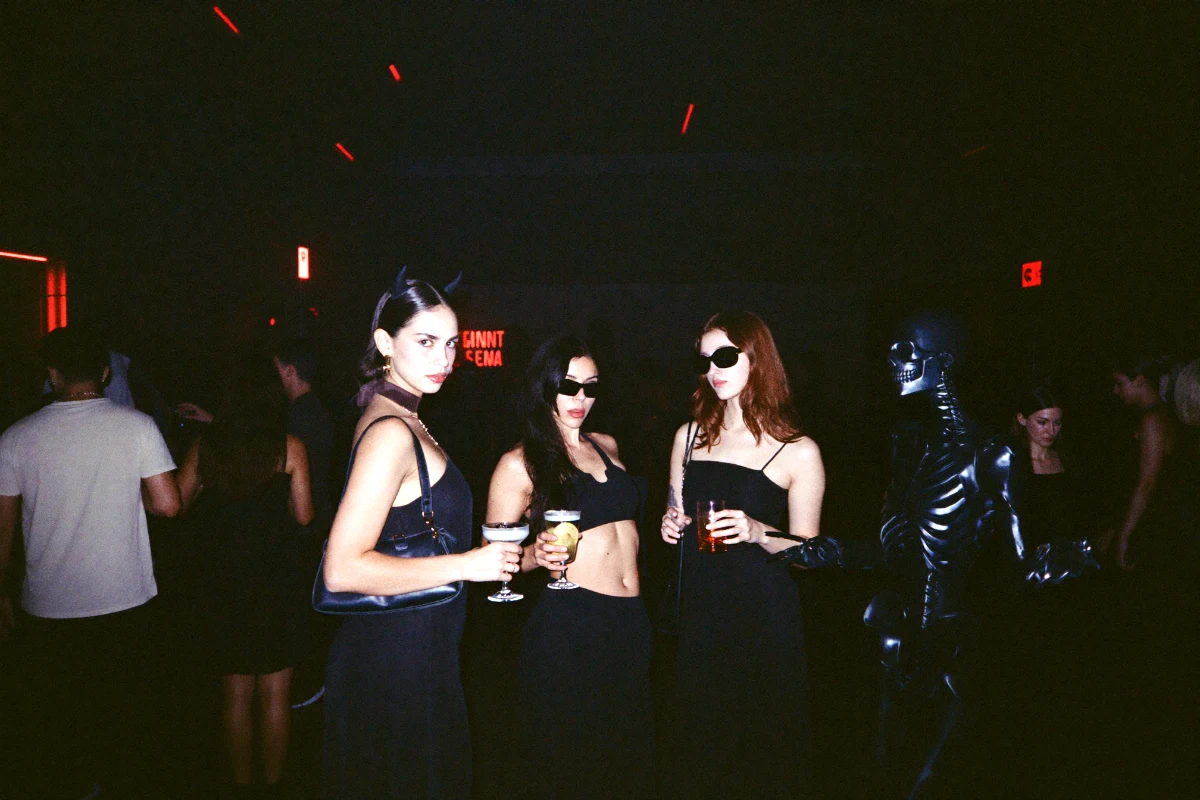 Friends in Halloween costumes with devil horns and a skeleton bodysuit holding cocktails at a warehouse party during a Halloween bar crawl