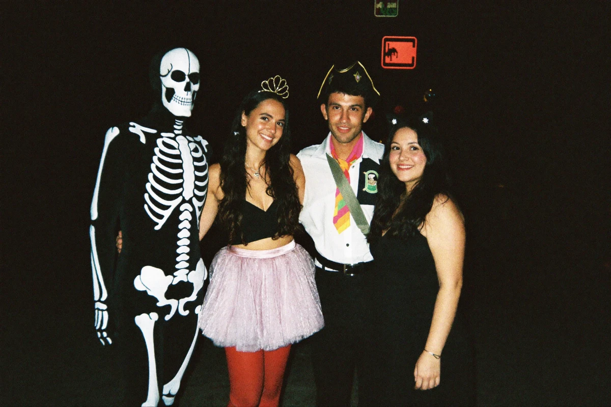 Four friends in Halloween costumes - a skeleton bodysuit, a tiara and pink tutu, a pirate hat and cat ears - staying together as a group outside a bar at night