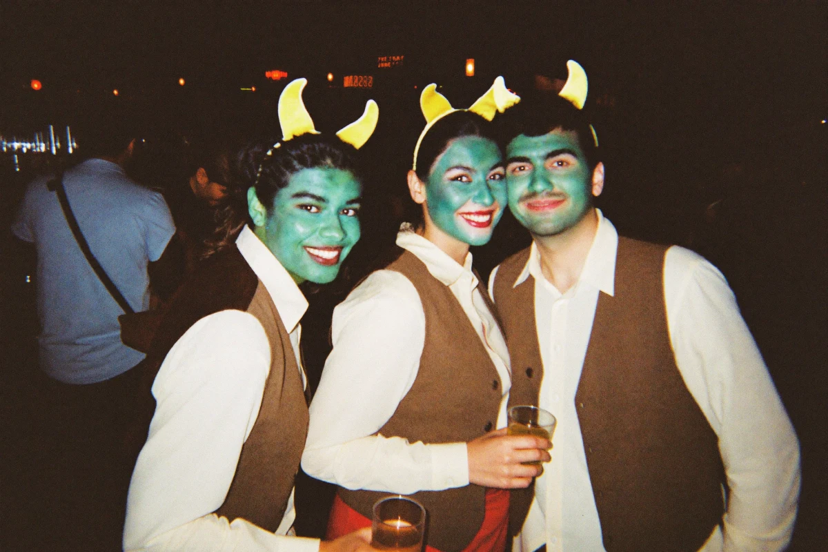 Three friends in matching ogre group costumes with green face paint, yellow ogre ear headbands and brown felt vests over cream shirts at a Halloween bar crawl