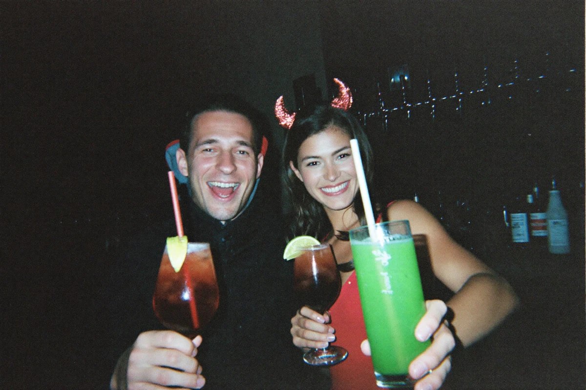 Two friends in Halloween costumes with red glitter devil horns holding up a blood-red cocktail and a glowing green cocktail at a dark bar