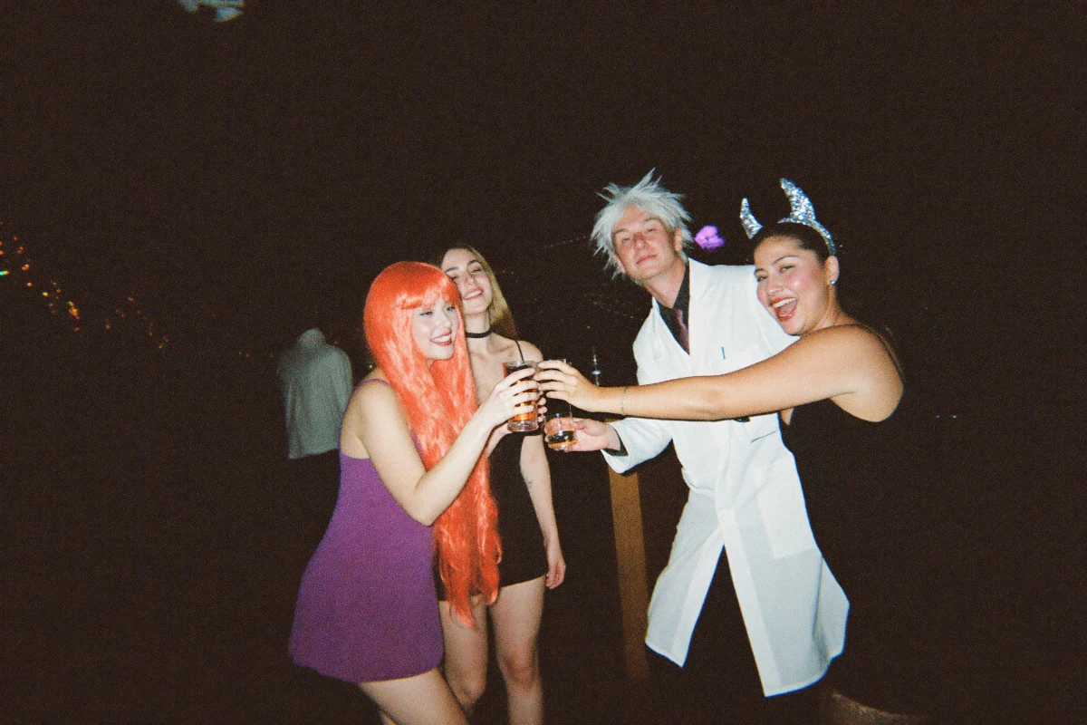 A group of friends in Halloween costumes including a long orange wig, a mad scientist lab coat with spiky white wig and glitter devil horns clinking glasses in a toast at a bar