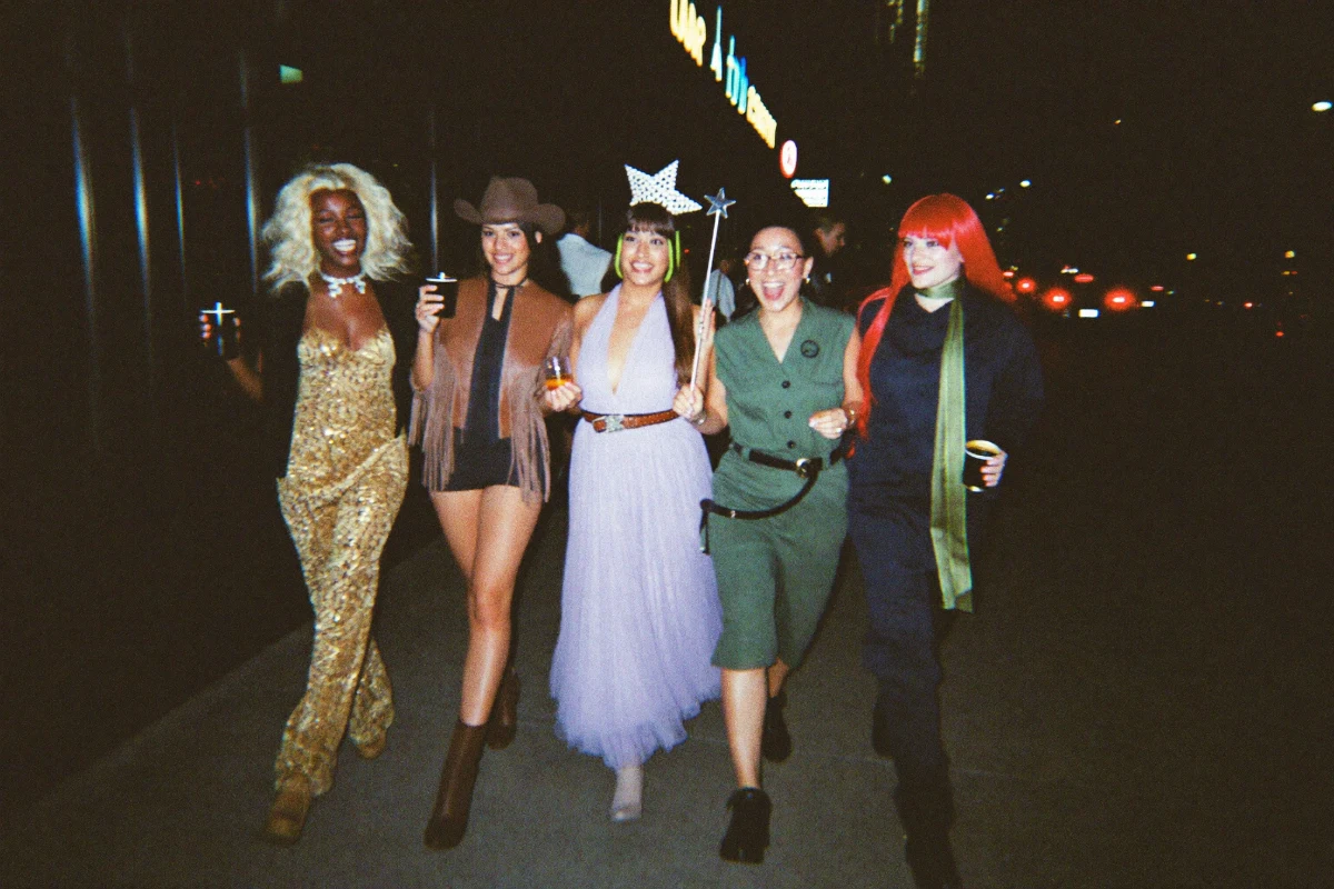 A group of friends in Halloween costumes including a disco jumpsuit, a cowboy hat and a tiara walking down a busy city sidewalk between bars at night