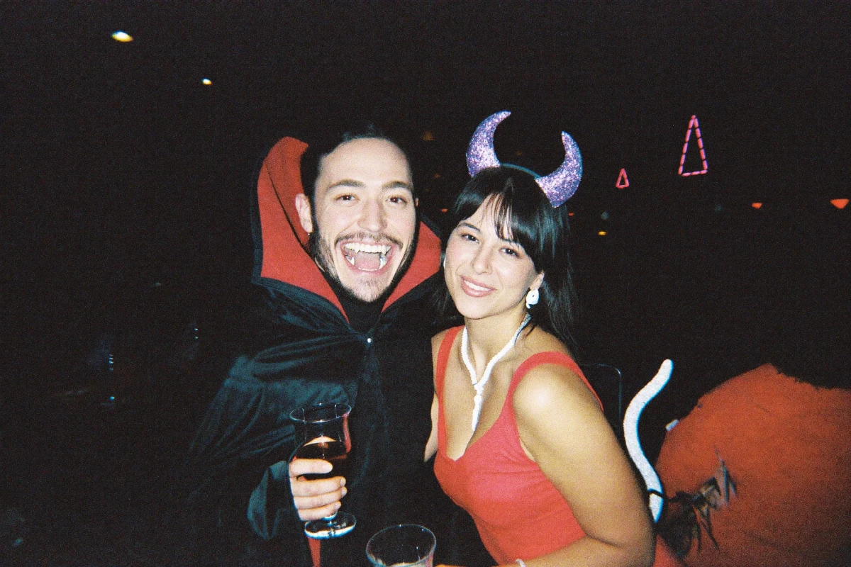 A couple in Halloween costumes at a crowded bar - a man in a black high-collared vampire cape with fangs beside a woman in a red dress with glitter devil horns and a tail, both holding drinks and smiling