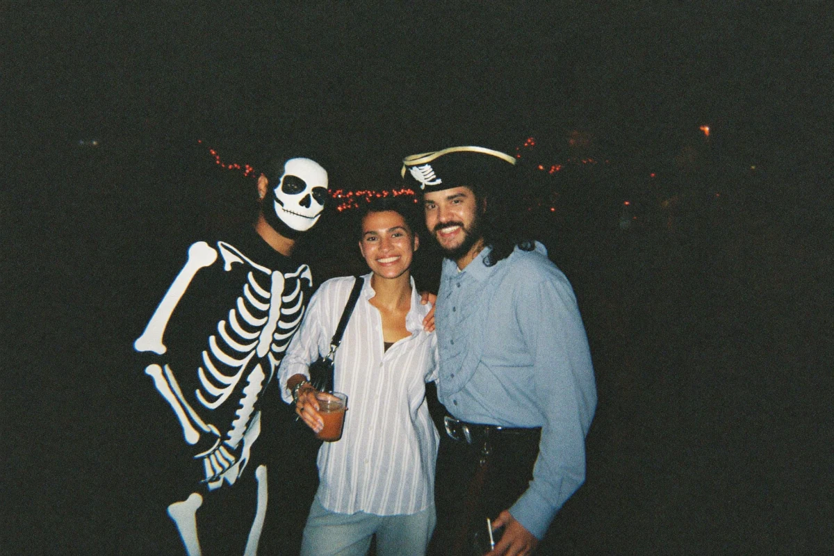 Two friends posing for a flash photo at a Halloween bar crawl, one in a skeleton costume with white skull face paint, the other in a pirate tricorn hat and blue shirt