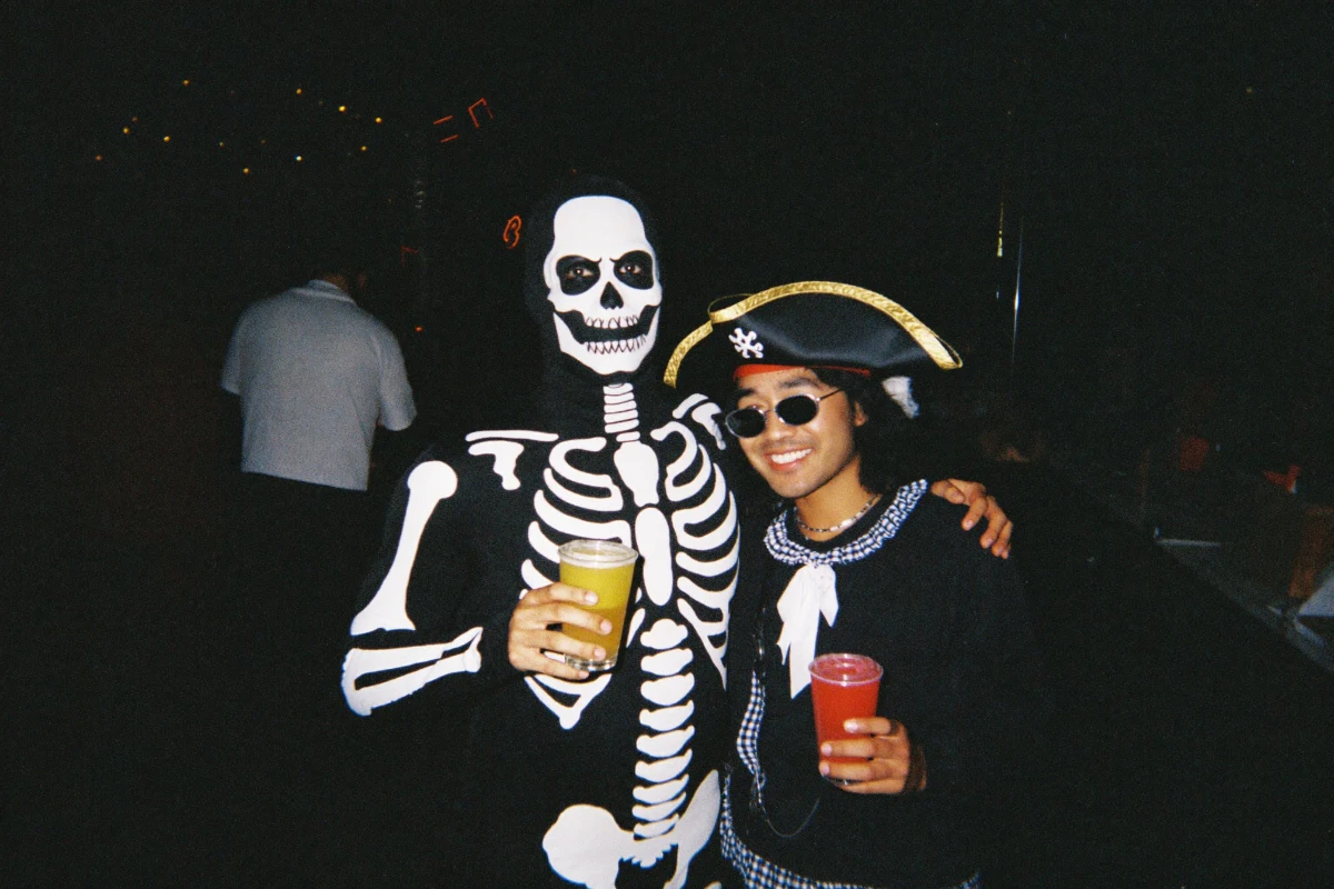 Two friends at a Halloween bar crawl, one in a black skeleton-print bodysuit with skull face paint and one in a pirate tricorn hat with a ruffled collar, smiling and holding drinks under camera flash