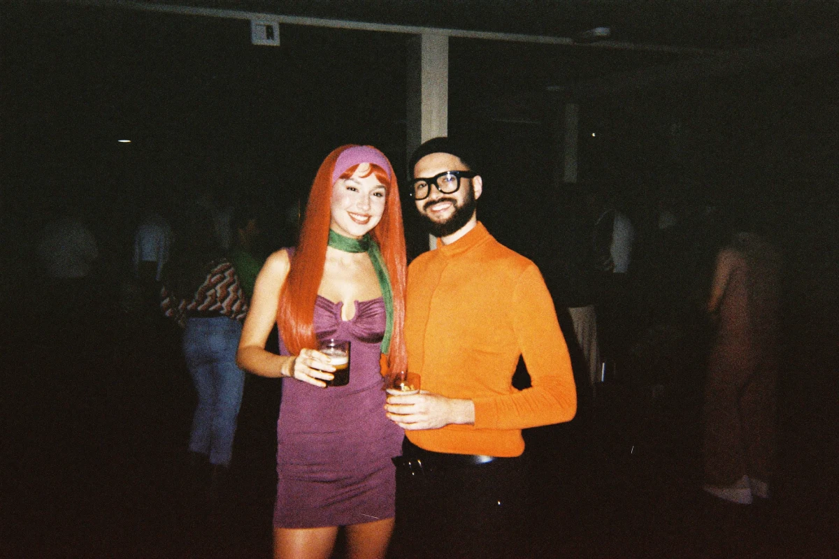 Two friends in a retro cartoon sleuth group costume at a Halloween bar crawl, one in a long orange wig, purple mini dress, purple headband and green neck scarf, the other in black-framed glasses and an orange turtleneck, smiling with drinks under camera flash