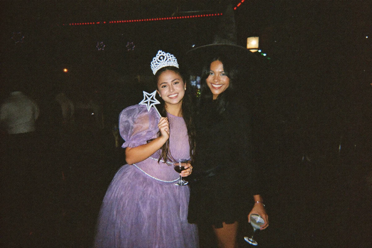 Two friends smiling at a Halloween bar, one in a lilac tulle fairy godmother costume with a tiara and star wand, the other in a black witch hat and dress