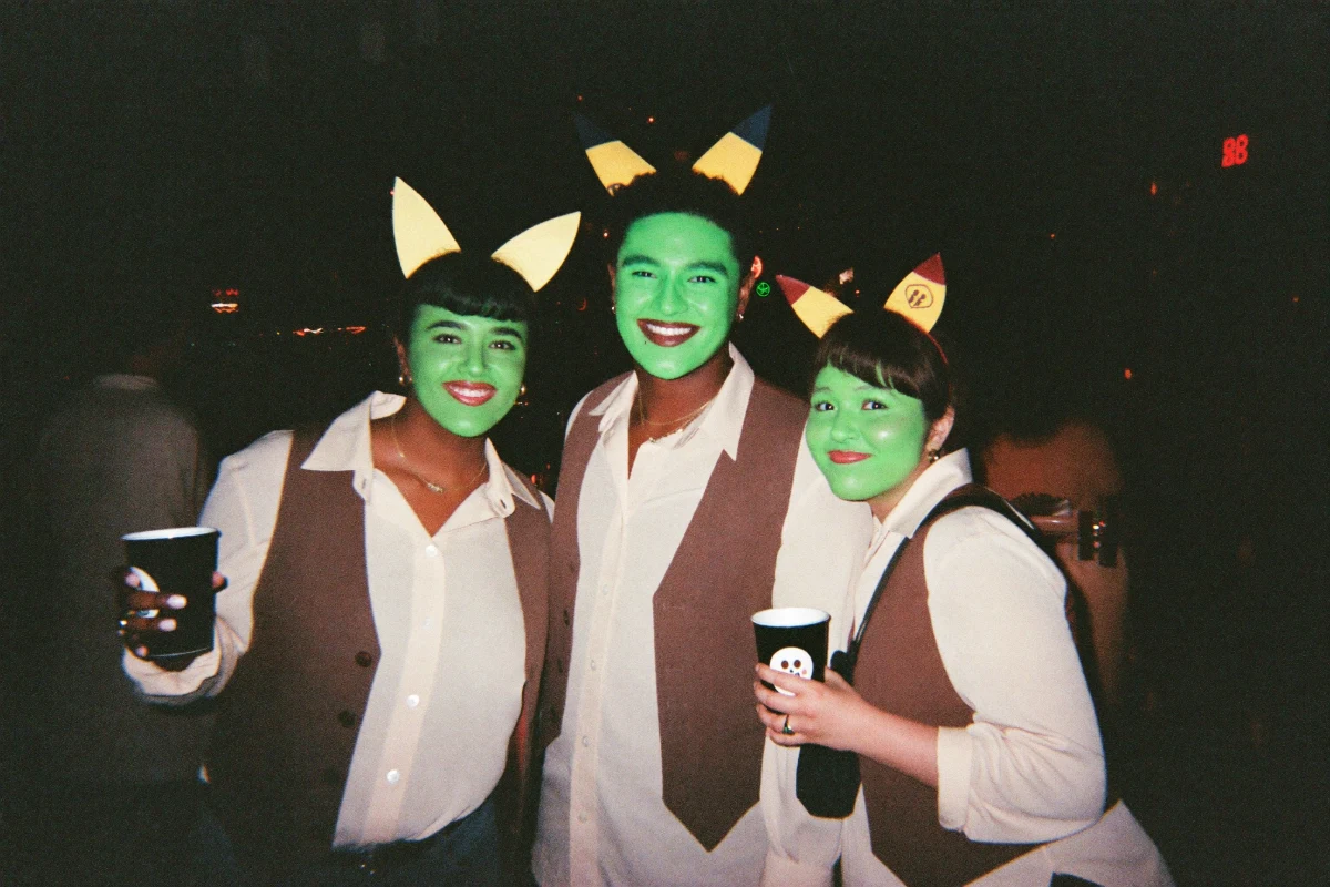 Three smiling friends in matching ogre costumes with green face paint, yellow ogre-ear headbands and brown felt vests holding drinks at a Halloween bar crawl