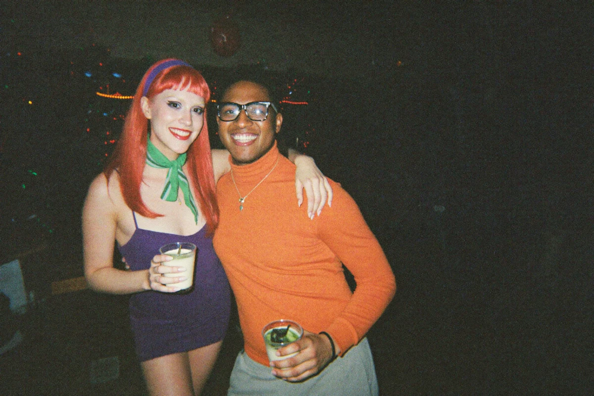 Two smiling friends posing at a Halloween bar crawl, one in a long orange wig, purple dress and green scarf and the other in thick black glasses and an orange turtleneck