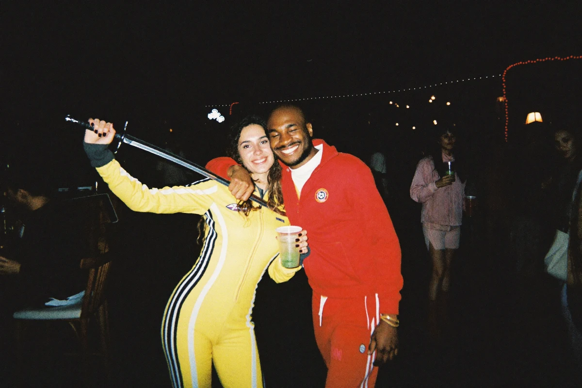 Two smiling friends at a Halloween bar crawl, one in a yellow motorcycle jumpsuit with black stripes holding a prop sword, the other in a red tracksuit holding a drink