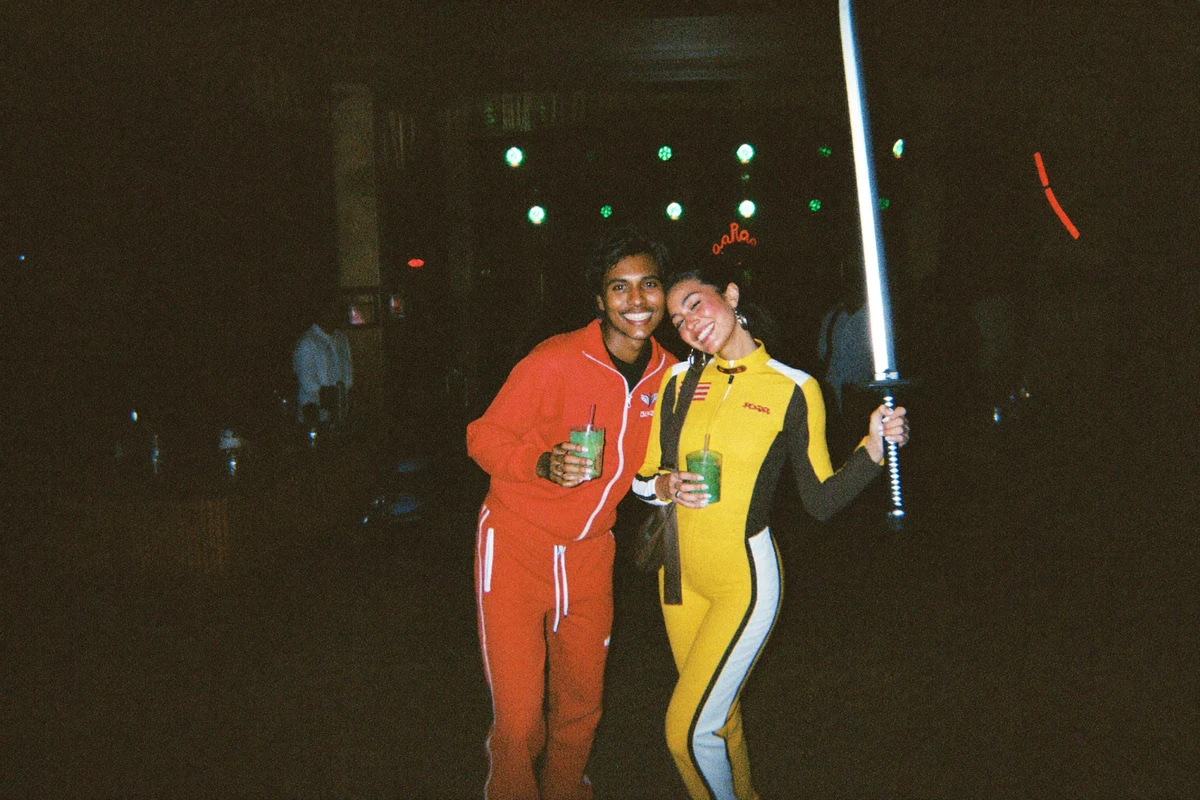 Two friends smiling in a dark bar, one in a yellow motorcycle jumpsuit holding a prop samurai sword and one in a red tracksuit, Halloween costume bar crawl