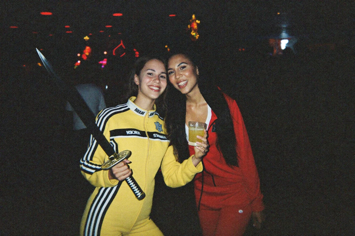 Two friends smiling at a Halloween bar crawl, one in a yellow motorcycle jumpsuit holding a prop samurai sword and one in a red tracksuit