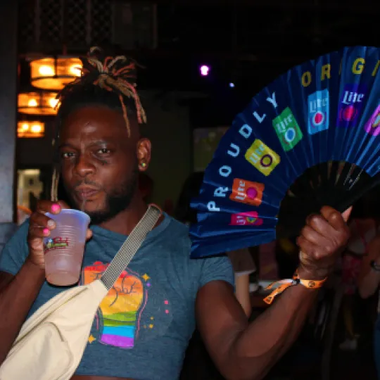 Young gentleman sips drink iconically and is holding his Pride fan during the Pride Bar Crawl