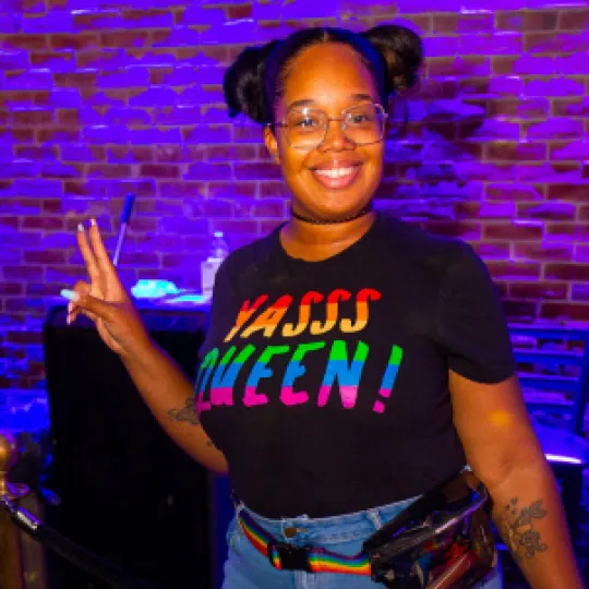 Young girl having the time of her life at the Pride bar crawl in Boston wearing a shirt that says "Yass Queen" and throwing up a peace sign.
