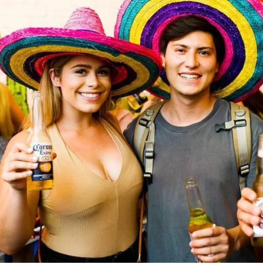 Vibrant young man and woman in eye-catching sombreros, dancing and relishing the energetic vibe of the Tacos and Tequila event while cheersing a festive corona beer
