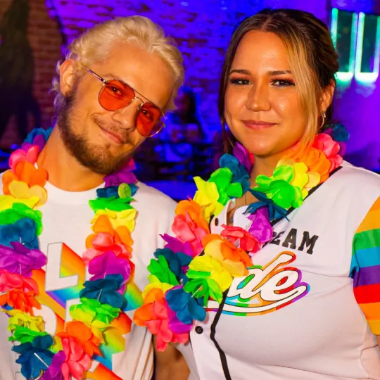 A guy and a girl smiling happily as they show off their rainbow lays and jerseys that read "Team Pride" at the Pride Bar Crawl