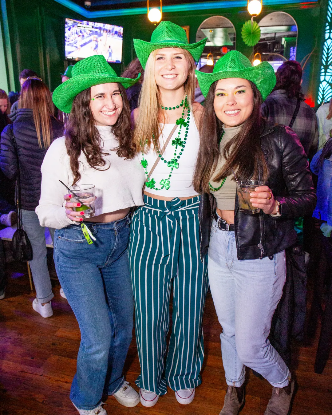 Beaming faces beneath green cowboy hats, a group of 3 girls smile in the vibrant ambiance of the bar crawl's st pats celebration
