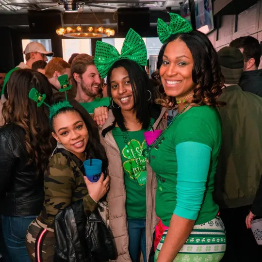 Ebullient friends decked out in green outfits echoing Irish pride, weaving through the bar crowd, leaving a trail of laughter and joy

