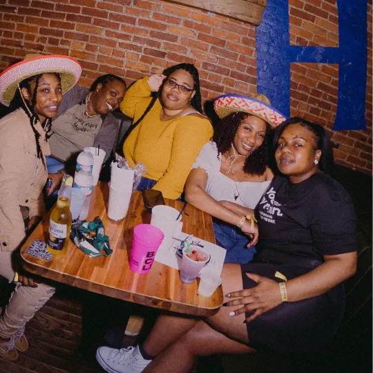 Happy and content group of friends gather around a table filled with tacos and tequila wearing sombreros showing with their laughter, and savoring every sip and bite the Tacos and Tequila night has to offer.
