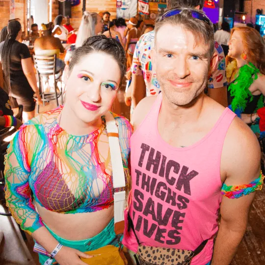 Friends pose to the camera during the Pride Bar Crawl and man shows a little comic relief wearing a tank saying "Thick Thighs Save Lives"