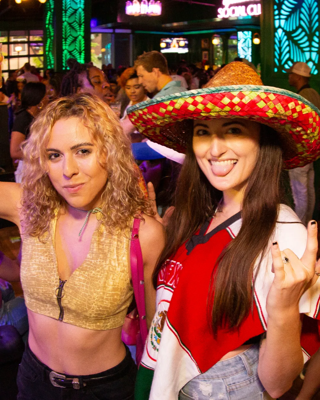 Animated duo, decked out in traditional cinco de mayo attire sharing laughs and toasts amidst the bustling bar crawl festivities
