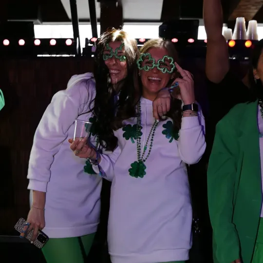 Group of jovial girlfriends united in their green shamrock glasses smiling playfully, sharing stories and drinks amidst the St. Patrick's Day bar crawl hustle

