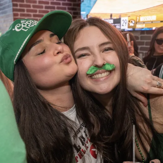 A girl in a green hat and a girl with a fake green mustache having fun during the St Patricks Bar Crawl 