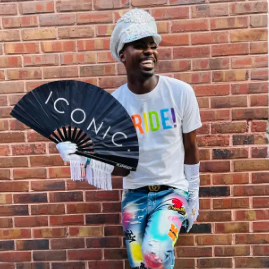 Young man posing outside of one of the best bars in Philly during the Pride Bar Crawl dressed in his most colorful pants and holding a Pride fan that says "Iconic"