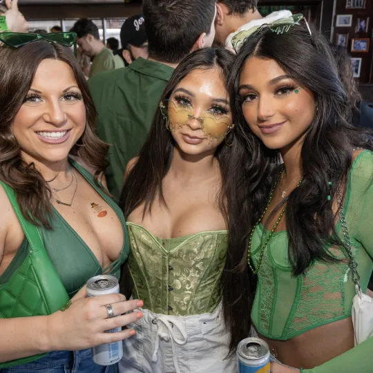 Animated young women decked out in traditional St. Patrick's attire, sharing laughs and toasts amidst the bustling bar crawl festivities.
