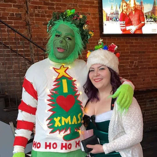 Man dressed in "The Grinch" makeup and outfit standing next to a girl dressed in an awesome christmas sweater getting ready for the ugly sweater bar crawl