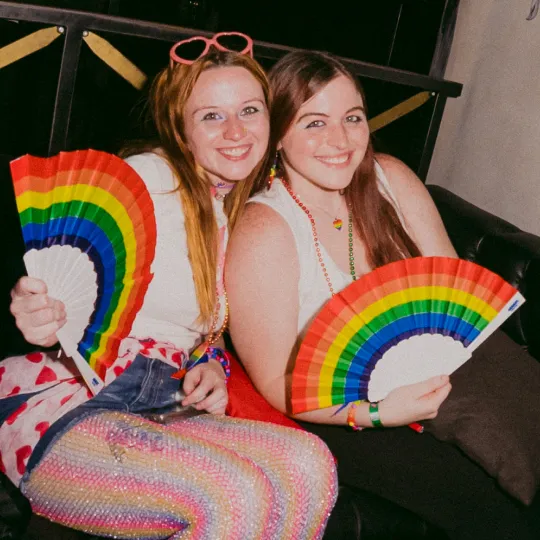 2 friends happy and living their best lives decked out in their most rainbow outfits and showcasing their rainbow Pride fans