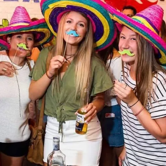 Snapshot of jubilant ladies, their wide-brimmed sombreros and costume mustaches casting playful shadows, as they enjoy the night's taco and tequila offerings during the bar crawl
