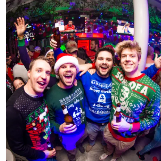 A cheerful group of 4 young gentleman bar-crawlers in ugly sweater attire, laughing and toasting with festive drinks in hand with a vibrant dance floor as the backdrop in one of the best nightclubs in Raleigh for the Ugly Sweater Bar Crawl