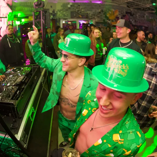 Beaming faces beneath green bucket hats hats and shamrock suits bask in the vibrant ambiance of the bar crawl's Irish celebration at the DJ booth
