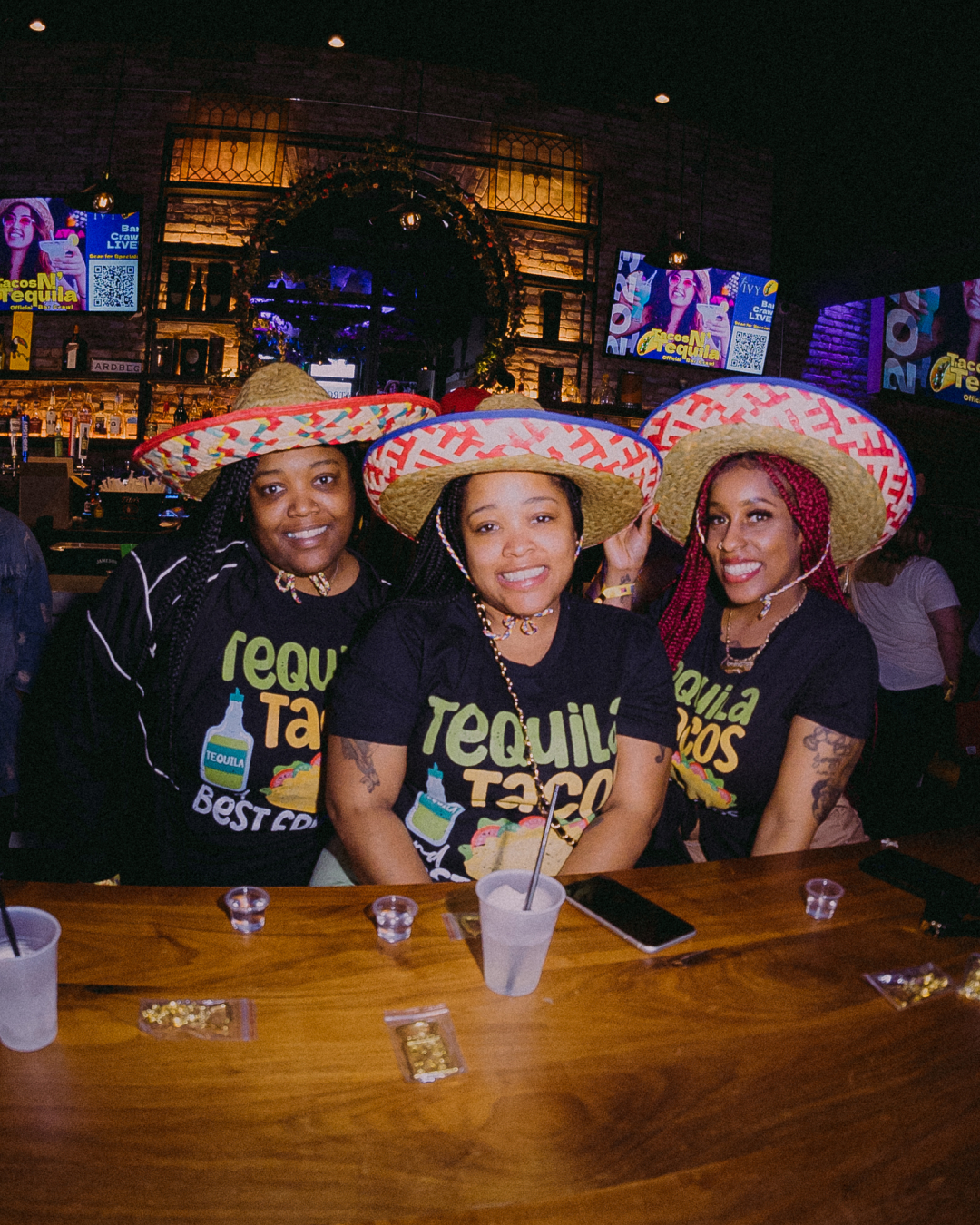 3 girls in sombrero hats sitting at a bar getting ready to take a tequila shot during the tacos and tequila bar crawl wearing a tshirt that says "Tequila Tacos and Best Friends"