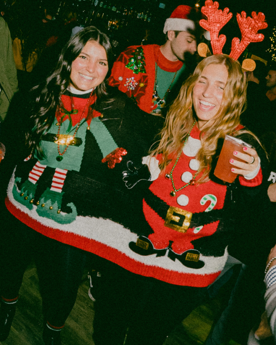 2 girls sharing one christmas sweater cheersing the camera during the holiday bar crawl
